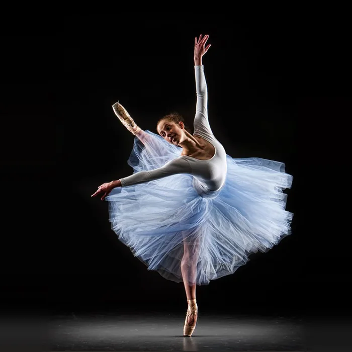 Ballet dancer in a white leotard and blue romantic tutu holding an arabesque en pointe on stage